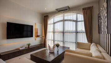 A modern living room with a flat-screen TV, a sofa set, a coffee table with decorative items, and a large window with curtains.