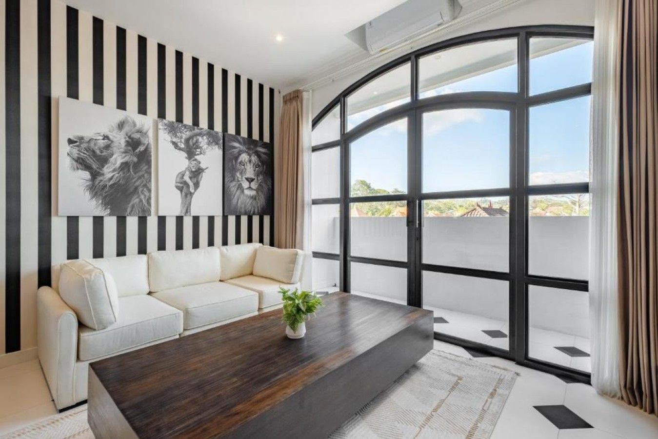 A modern living room with a white sofa, a dark wooden coffee table, and large windows offering a view of the outside.