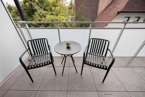 Ein Balkon mit zwei Stühlen und einem kleinen Tisch, eine Topfpflanze auf dem Tisch und im Hintergrund eine Aussicht auf Bäume und Gebäude.