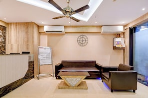 A modern lobby with a ceiling fan, air conditioning units, a wall clock, a sofa, a coffee table, and a decorative item on the wall. - Palette Dumdum Airport Formerly Seventy (Kolkata)