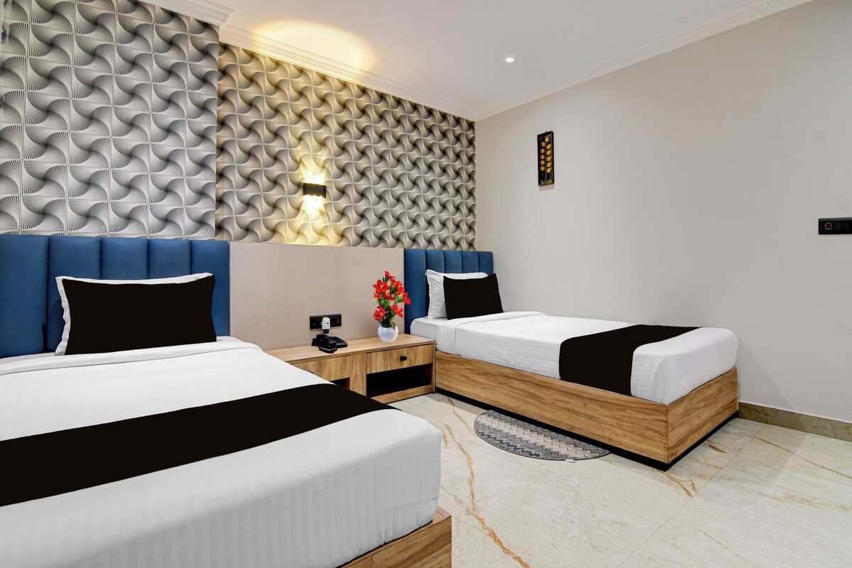 Habitación de hotel con dos camas, un papel tapiz estampado, una mesita de noche y una pequeña mesa con un jarrón de flores.
