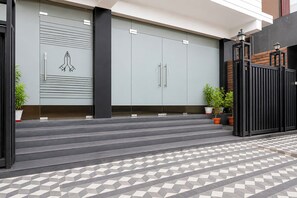Entrada moderna de un edificio con una serie de escalones que conducen a una zona vallada, flanqueada por plantas en macetas y un suelo de baldosas con diseño.