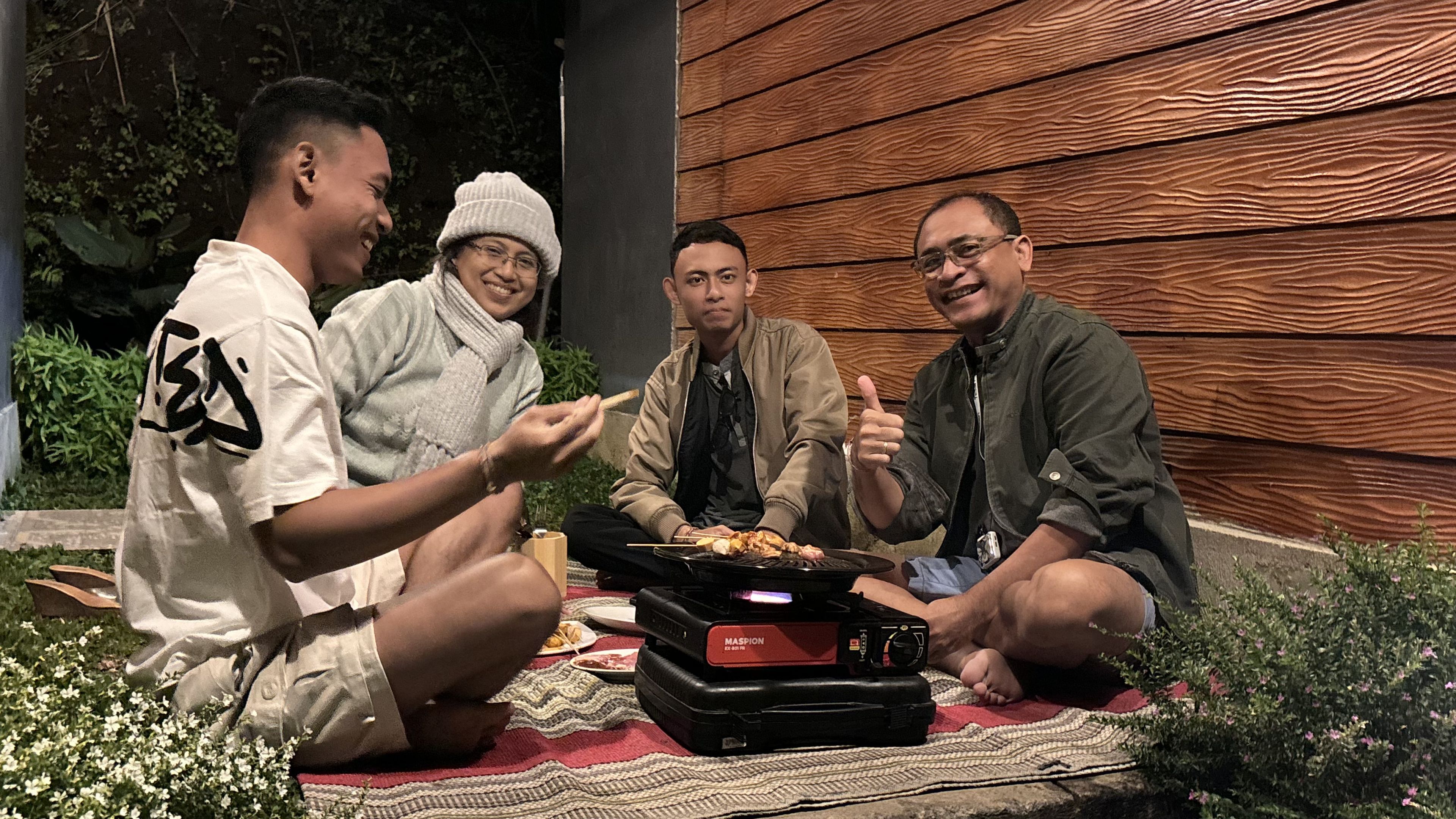 Four people sitting on a blanket outdoors with a portable grill.