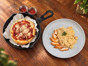 Une pizza garnie et une assiette de pâtes au poulet, le tout posé sur une table en bois.