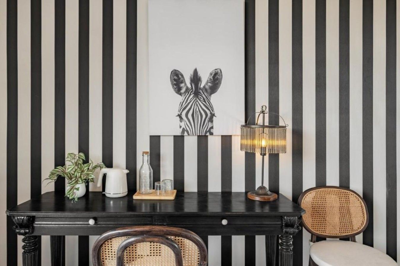 A room with a black and white striped wall, a zebra-themed artwork, a table with a lamp, a wicker chair, and a potted plant.