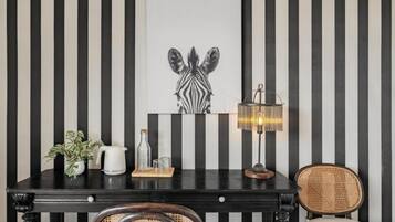 A room with a black and white striped wall, a zebra-themed artwork, a table with a lamp, a wicker chair, and a potted plant.