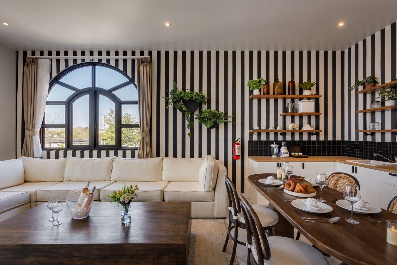 A modern living room with a large window, a sofa, a dining table with chairs, and a kitchen area with shelves and plants.