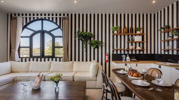 A modern living room with a large window, a sofa, a dining table with chairs, and a kitchen area with shelves and plants.