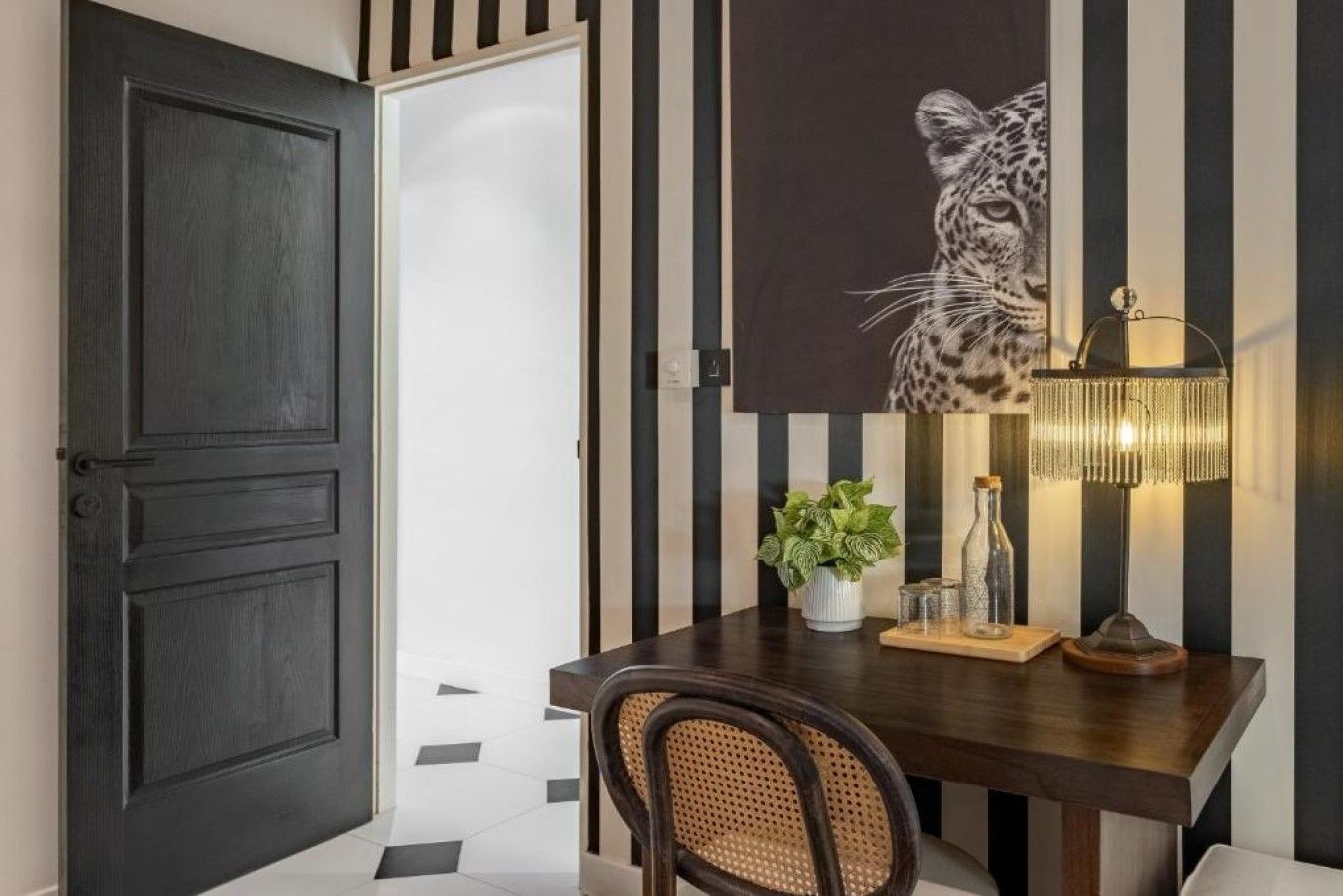 A room with a dark wooden door, a white wall with black stripes, a table with a plant, a lamp, and a chair.