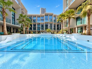 A modern hotel with a central pool, palm trees, and a clear sky. - Cozy Studio with Pool & Gym Near Circle Mall (Dubai)