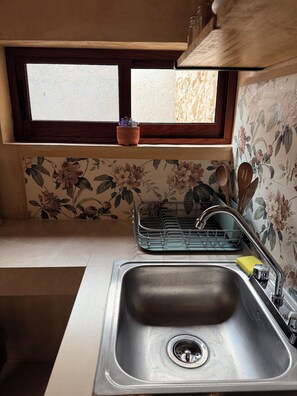 A kitchen with a stainless steel sink, a window with a potted plant, and floral wallpaper. - Las JULIAS Cuarto privado (Mérida)