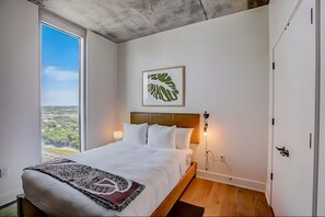 A bedroom with a bed, a view of the outdoors, a framed leaf artwork, and a wooden floor. - Rooftop Pool + Skyline Views • Near Rainey Street + Downtown | Panoramic Summit (Austin)