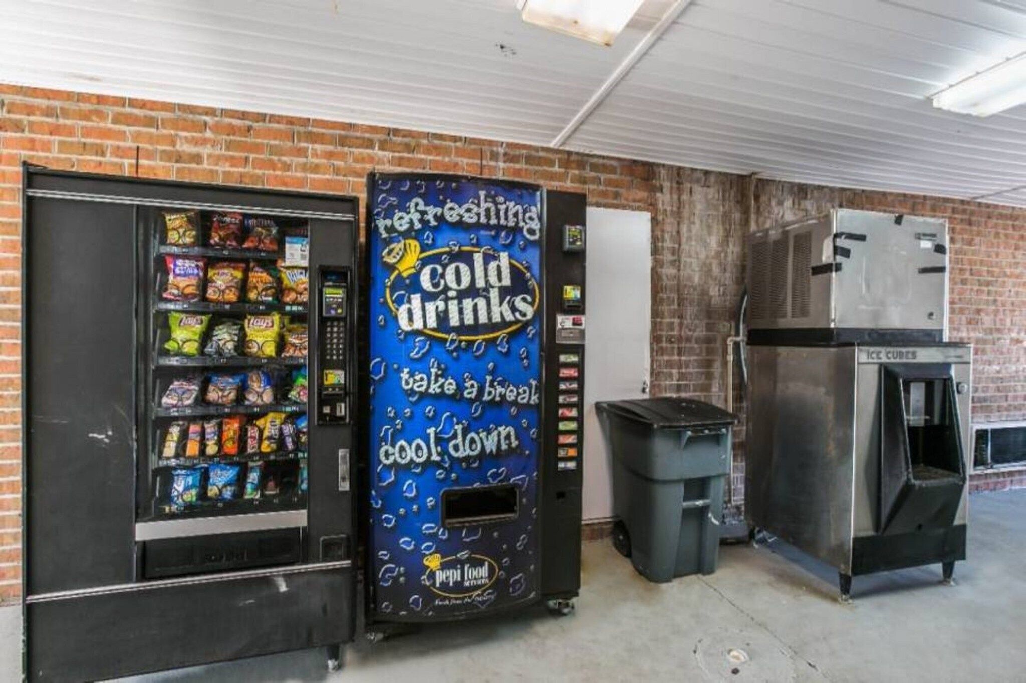 A vending machine with a blue exterior and a sign advertising cold drinks. Next to it is a trash bin and a refrigerator.