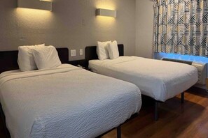 A hotel room with two beds, a window with patterned curtains, and a wall-mounted heater.