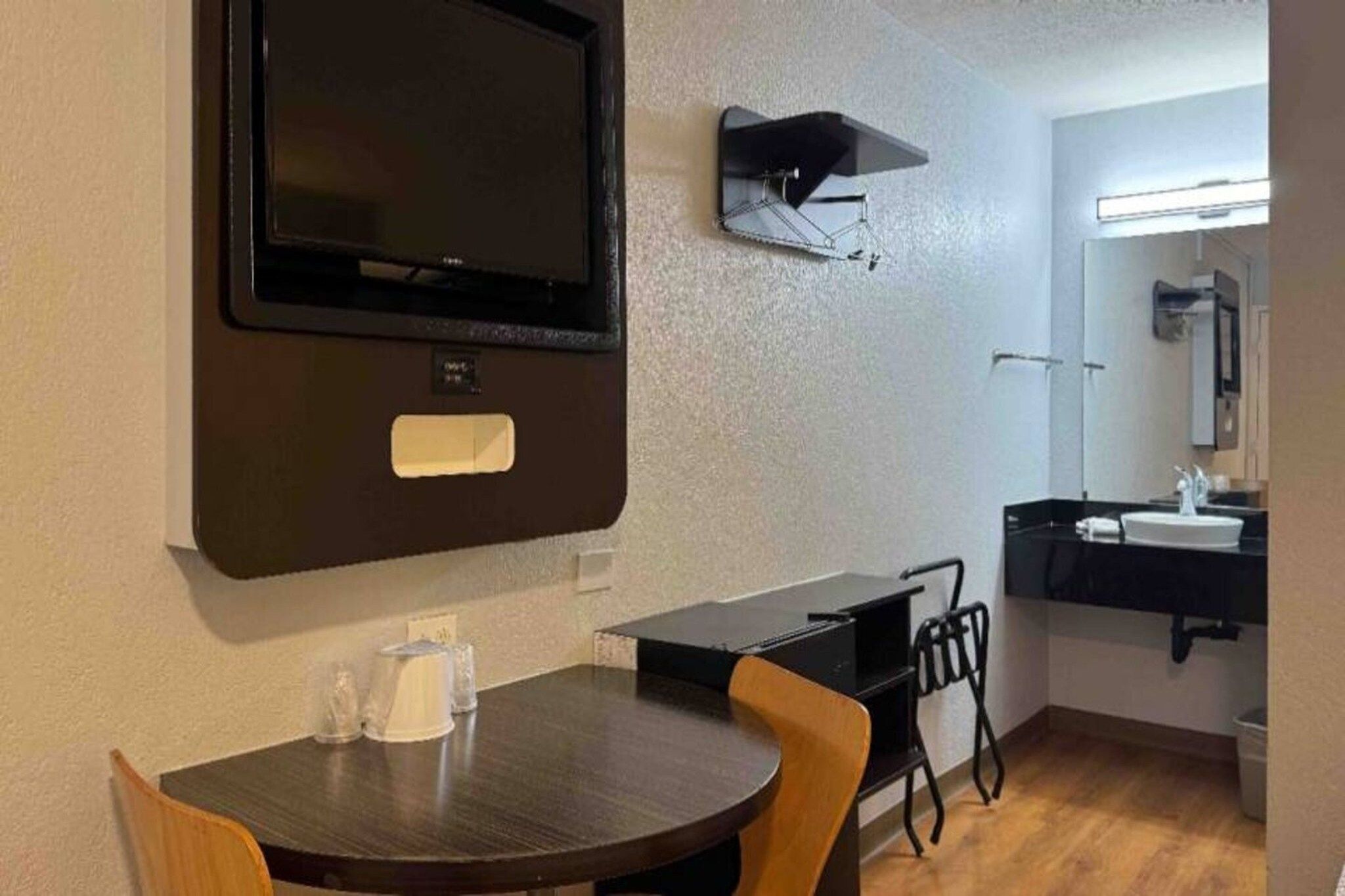 A compact room with a television mounted on the wall, a small table with chairs, a sink, and a mirror in the corner.