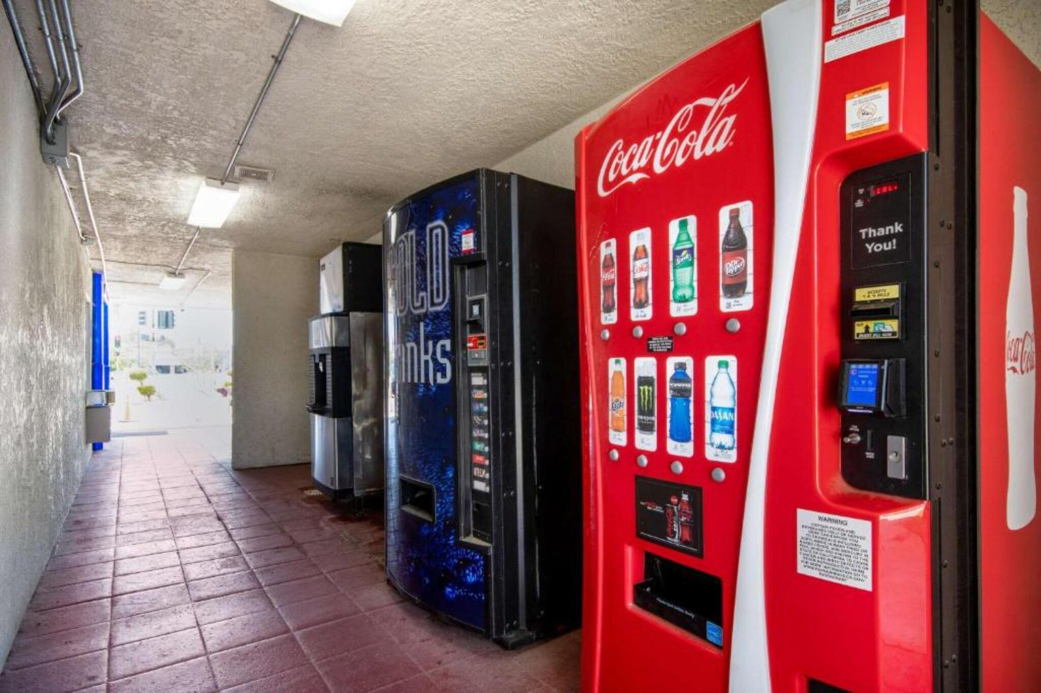 A row of vending machines with various beverage options, including Coca-Cola, in a corridor with tiled flooring.