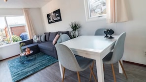 A modern living room with a sofa, a dining table with chairs, a potted plant, and a window with curtains. - DD 85 - Duindoorn Campground (IJmuiden)
