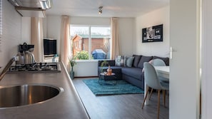 A modern kitchen with a stainless steel sink and stove, a dining area with a grey sofa and a small round table, and a window with curtains. - DD 85 - Duindoorn Campground (IJmuiden)