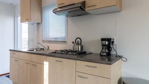 A kitchen with a gas stove, kettle, coffee maker, and a window with blinds. - DD 85 - Duindoorn Campground (IJmuiden)