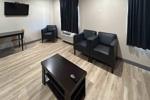 A room with a dark wooden table, two black armchairs, a small wooden chair, a standing heater, and a flat-screen TV mounted on the wall.
