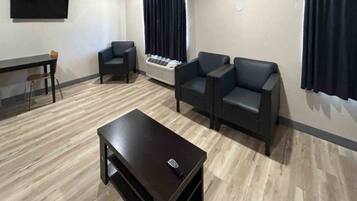 A room with a dark wooden table, two black armchairs, a small table with a remote, a standing heater, and a flat-screen TV mounted on the wall.