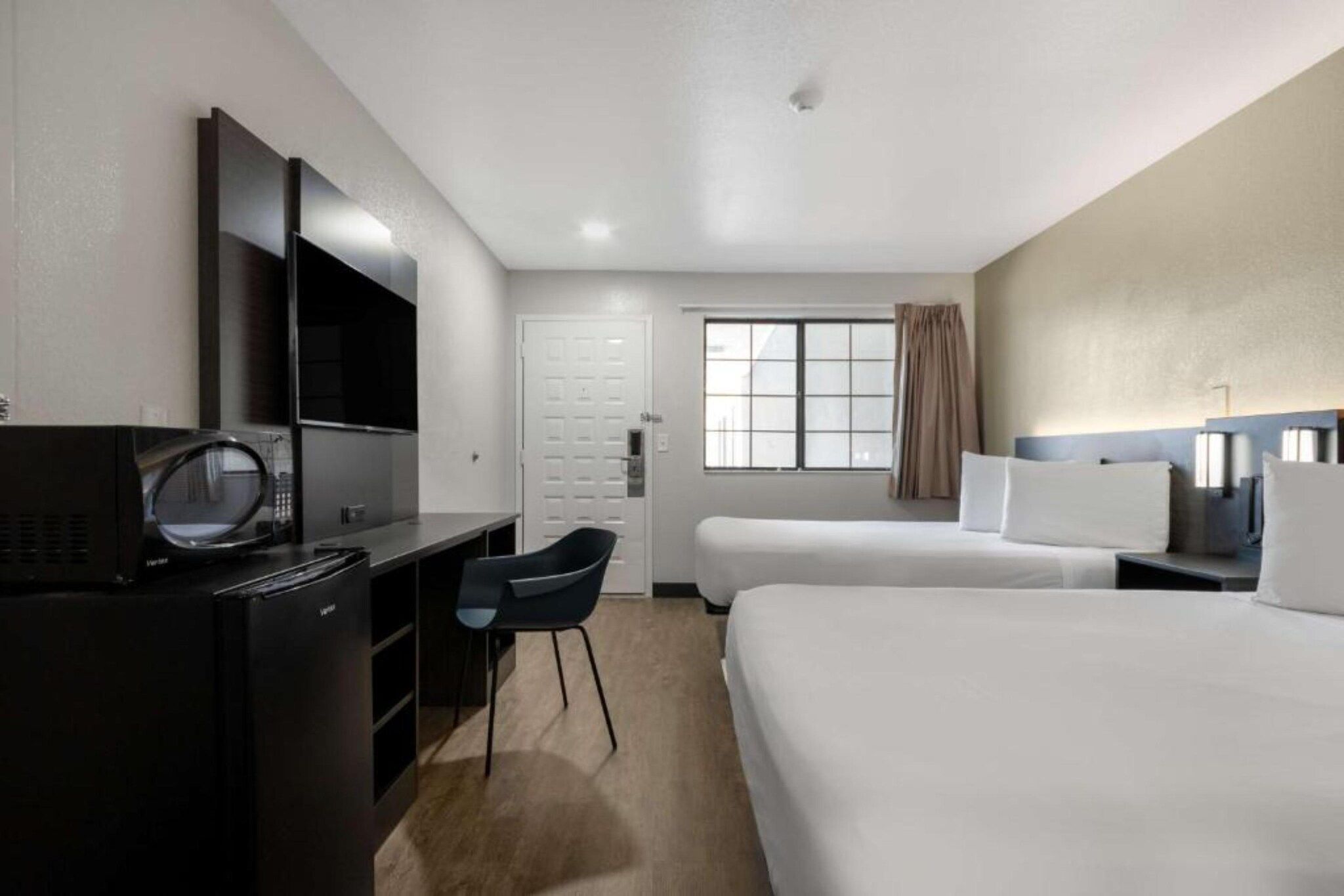 A hotel room with a desk, chair, two beds, a TV, and a window with curtains.
