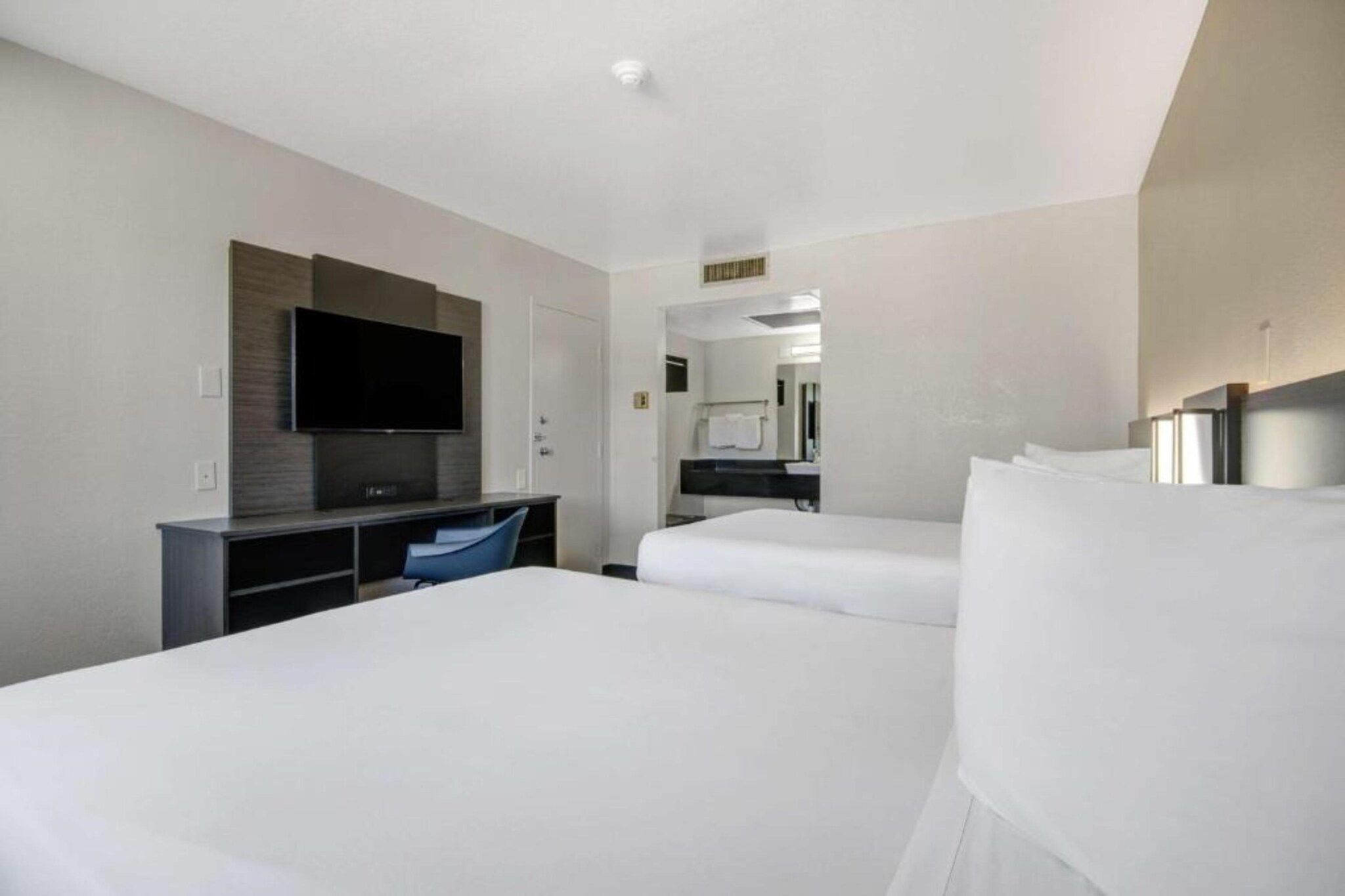 Quarto de hotel com duas camas, uma escrivaninha, televisão de tela plana e banheiro visível por uma porta aberta.