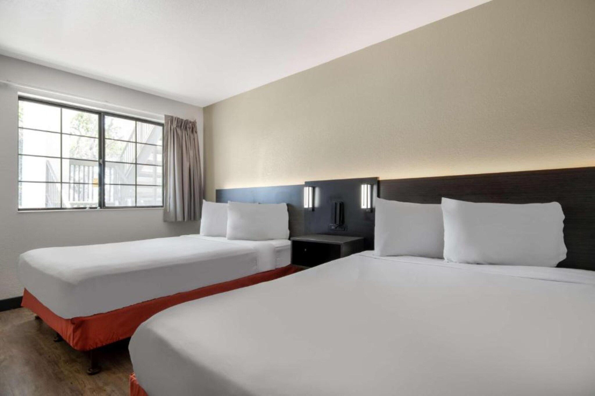 Quarto de hotel com duas camas, uma mesa de cabeceira, uma luminária e uma janela com cortinas.