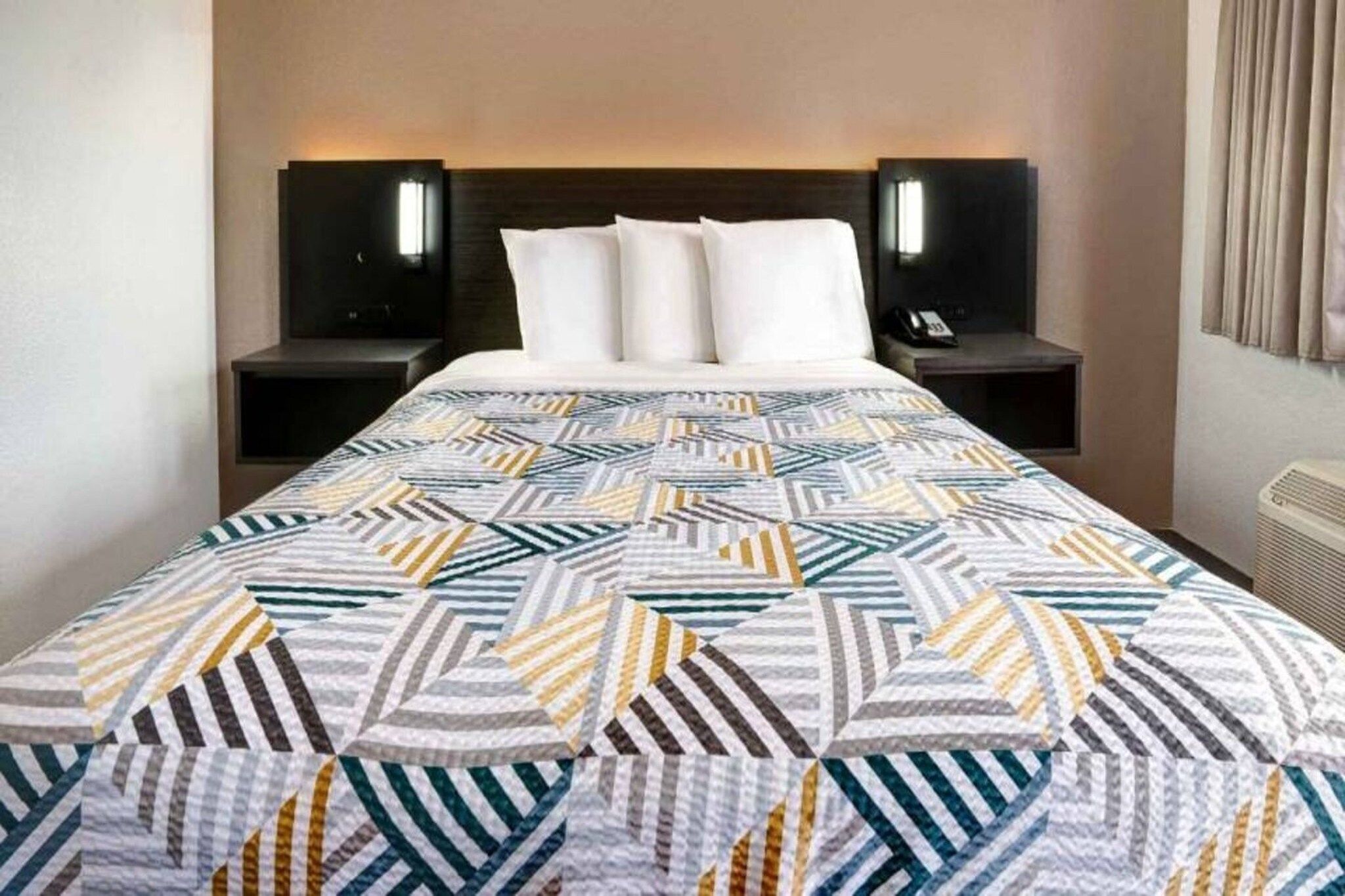 A hotel room with a bed, two bedside tables, and a wall-mounted air conditioning unit.