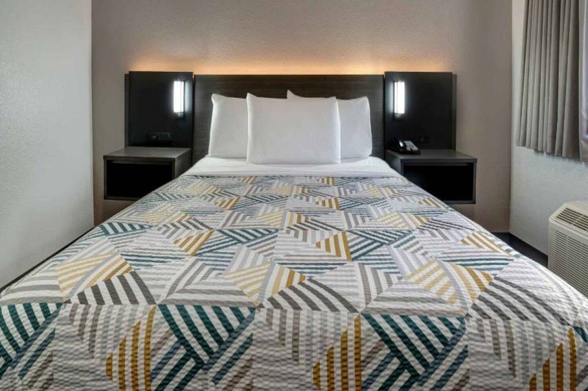 A hotel room with a bed, two bedside tables, and a wall-mounted air conditioning unit.