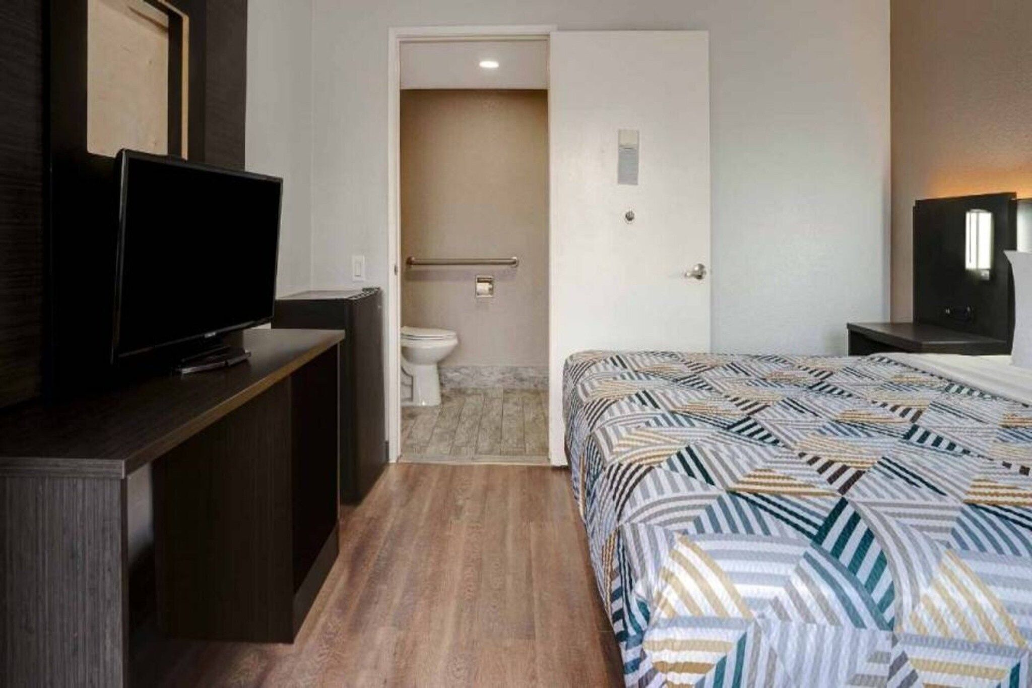 A hotel room with a bed, a television on a stand, a bathroom with a toilet and shower, and a door leading to another room.