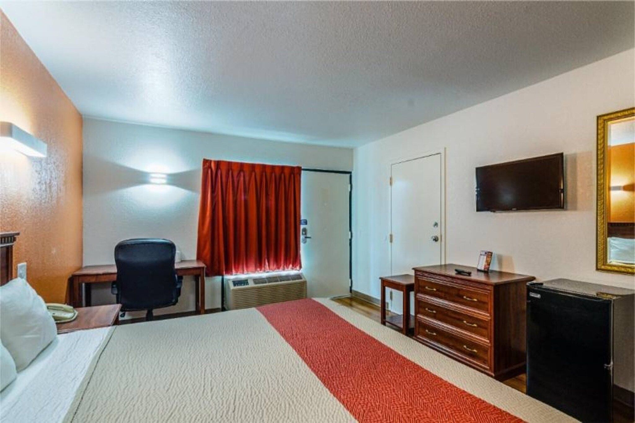 Ein Hotelzimmer mit einem Bett, einem Schreibtisch, einem Stuhl, einer Kommode, einem Fernseher und einem Fenster mit Vorhängen.