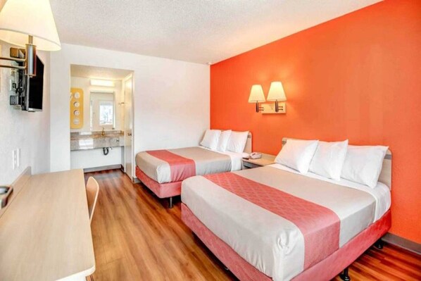 Habitación de hotel con dos camas, un escritorio, una silla, baño con espejo y una lámpara de pared.