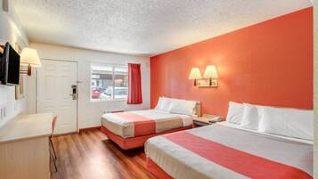A hotel room with two beds, a desk, a chair, and a window with a view of parked cars.