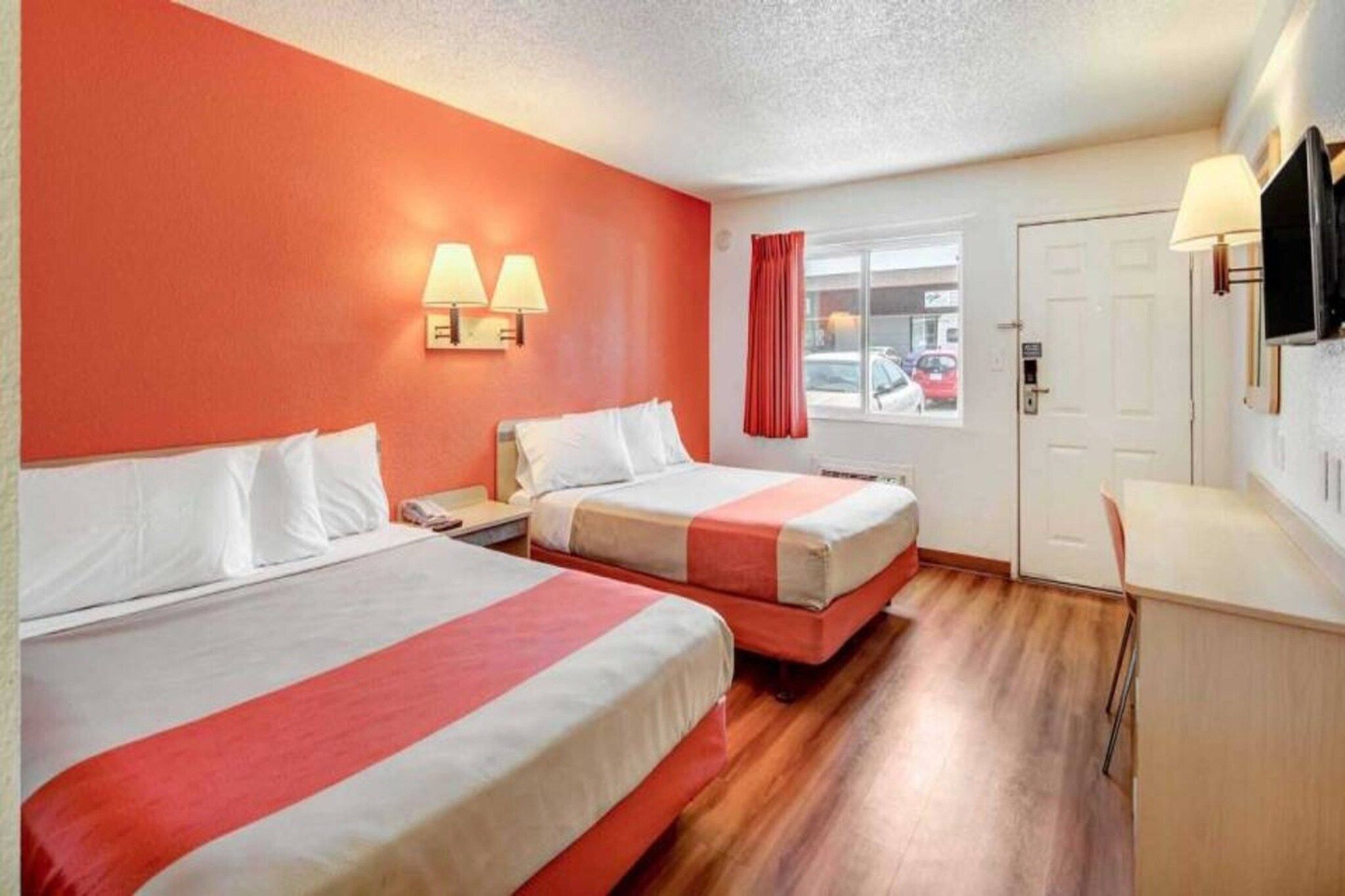 A hotel room with two beds, a desk, a TV, and a window with a view of the street.