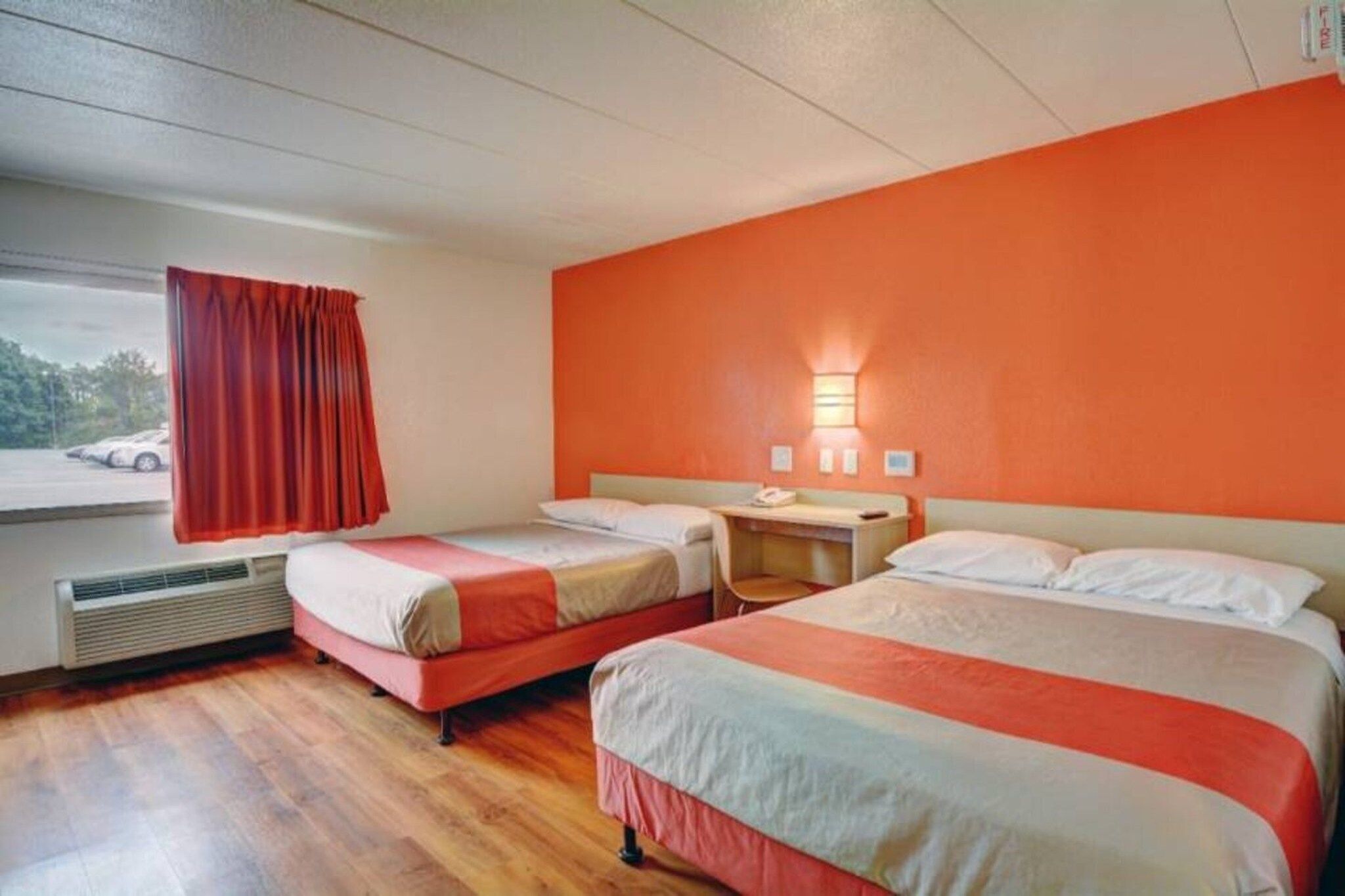 A hotel room with two beds, a desk, a window with curtains, and a wall-mounted light.