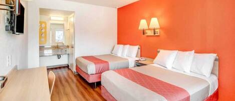 A hotel room with two beds, a desk, a chair, a bathroom with a mirror, and a wall-mounted lamp.