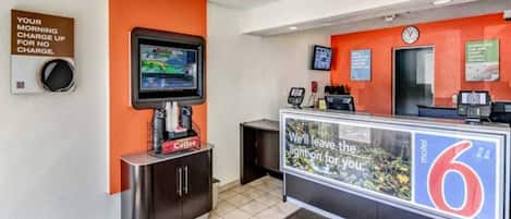 Uma área de recepção com tela digital, estação de café e balcão com placa.