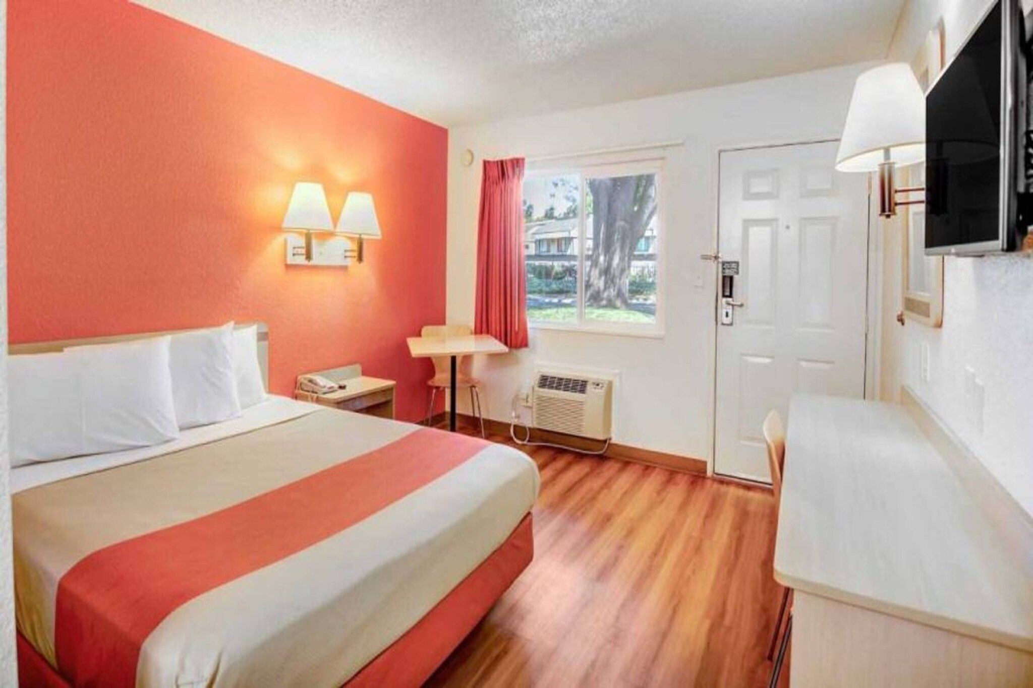 Ein Hotelzimmer mit Bett, Nachttisch, Lampe, Fenster und einem an der Wand befestigten Fernseher.