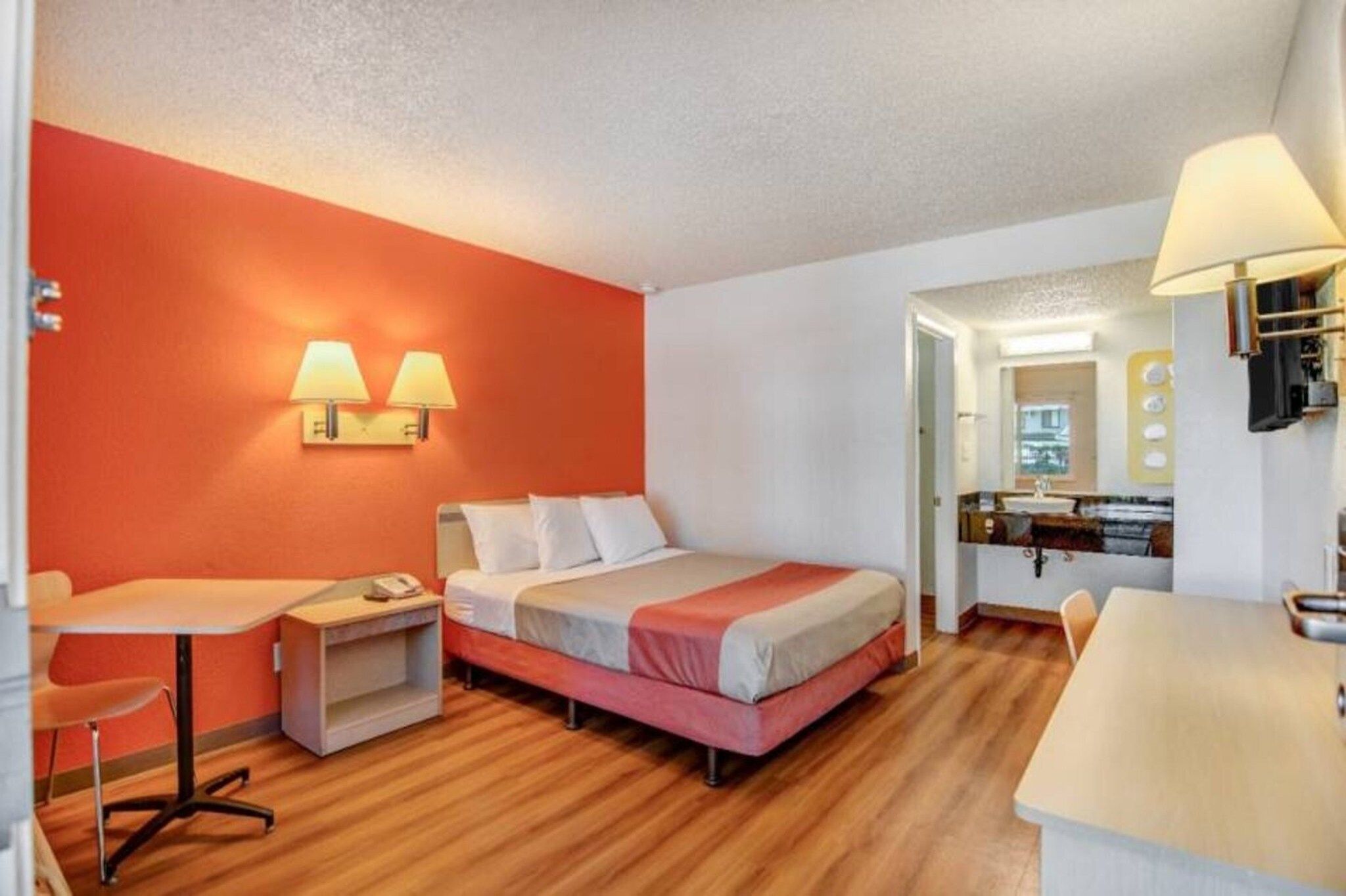 A hotel room with a bed, bedside table, lamp, and a small desk with a chair.