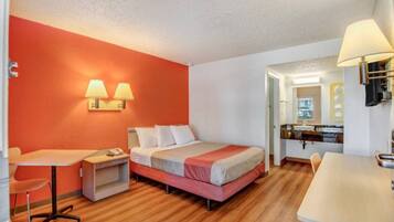A hotel room with a bed, bedside table, lamp, and a small desk with a chair.