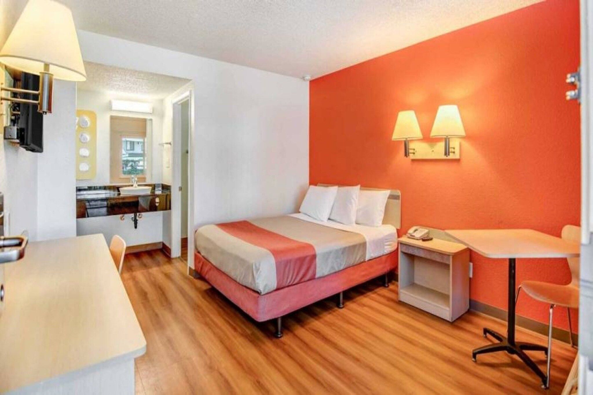 A hotel room with a bed, a desk, a chair, a small table, and a lamp.