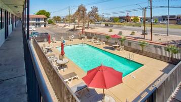 A swimming pool with lounge chairs and umbrellas, surrounded by a fence, with buildings in the background.