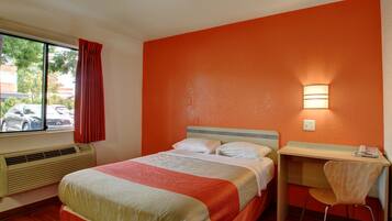 A hotel room with a bed, a desk, a chair, a window with a view of parked cars, and an air conditioning unit.