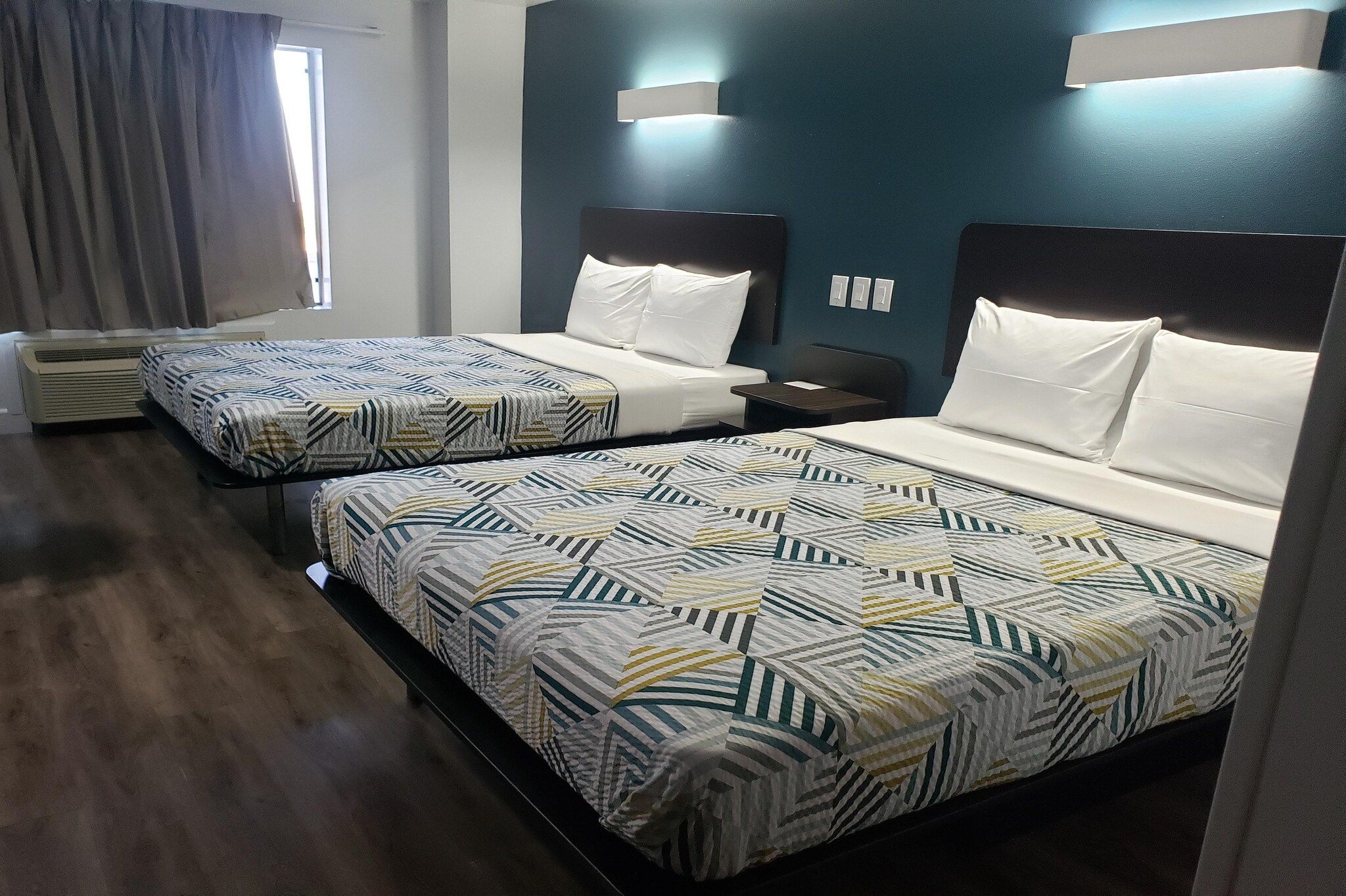 Hotellihuone, jossa on kaksi sänkyä, ikkuna verhoilla, seinälle asennettu ilmastointilaite ja yöpöytä.