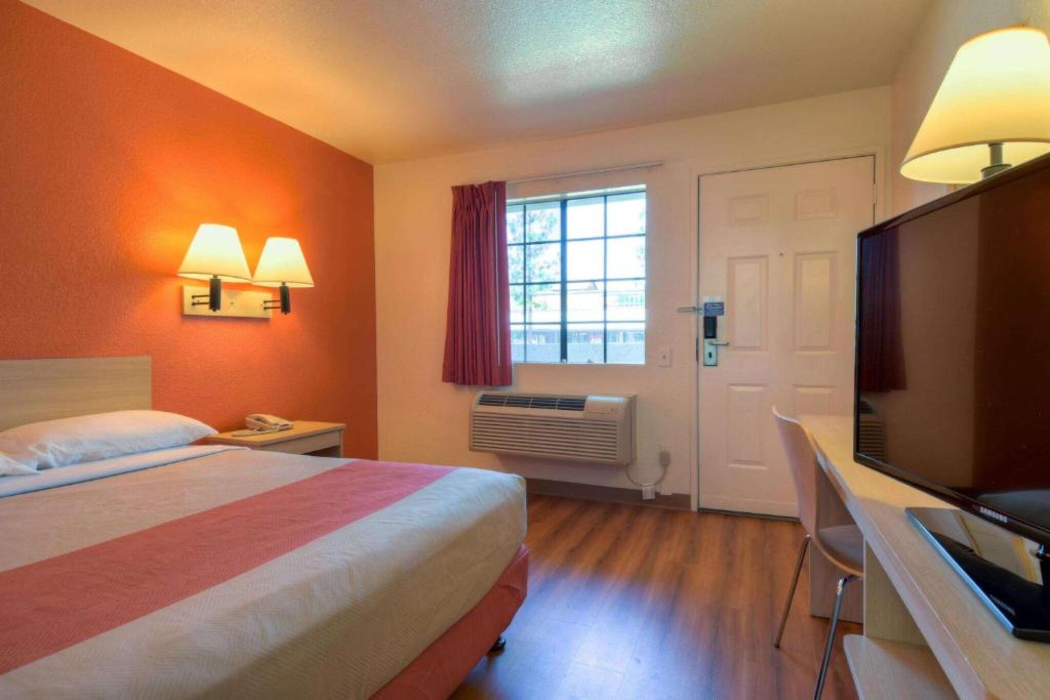 A hotel room with a bed, bedside lamps, a desk with a television, a window with curtains, and a door.