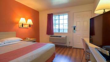 A hotel room with a bed, bedside lamps, a desk with a television, a window with curtains, and a door.