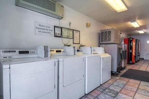 Une buanderie avec des lave-linge et des sèche-linge blancs, un panneau indiquant que « le motel 6 n’est pas responsable des objets laissés sans surveillance », et un distributeur automatique de Coca-Cola.