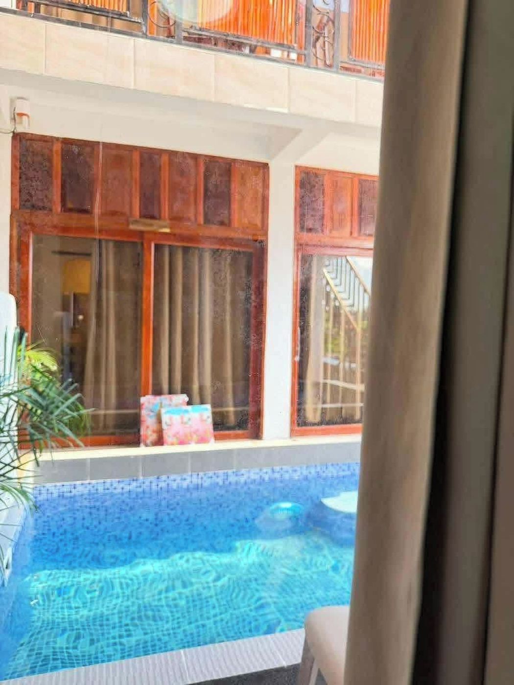 A swimming pool with a tiled deck, wooden window frames, and a view of a building with a balcony.