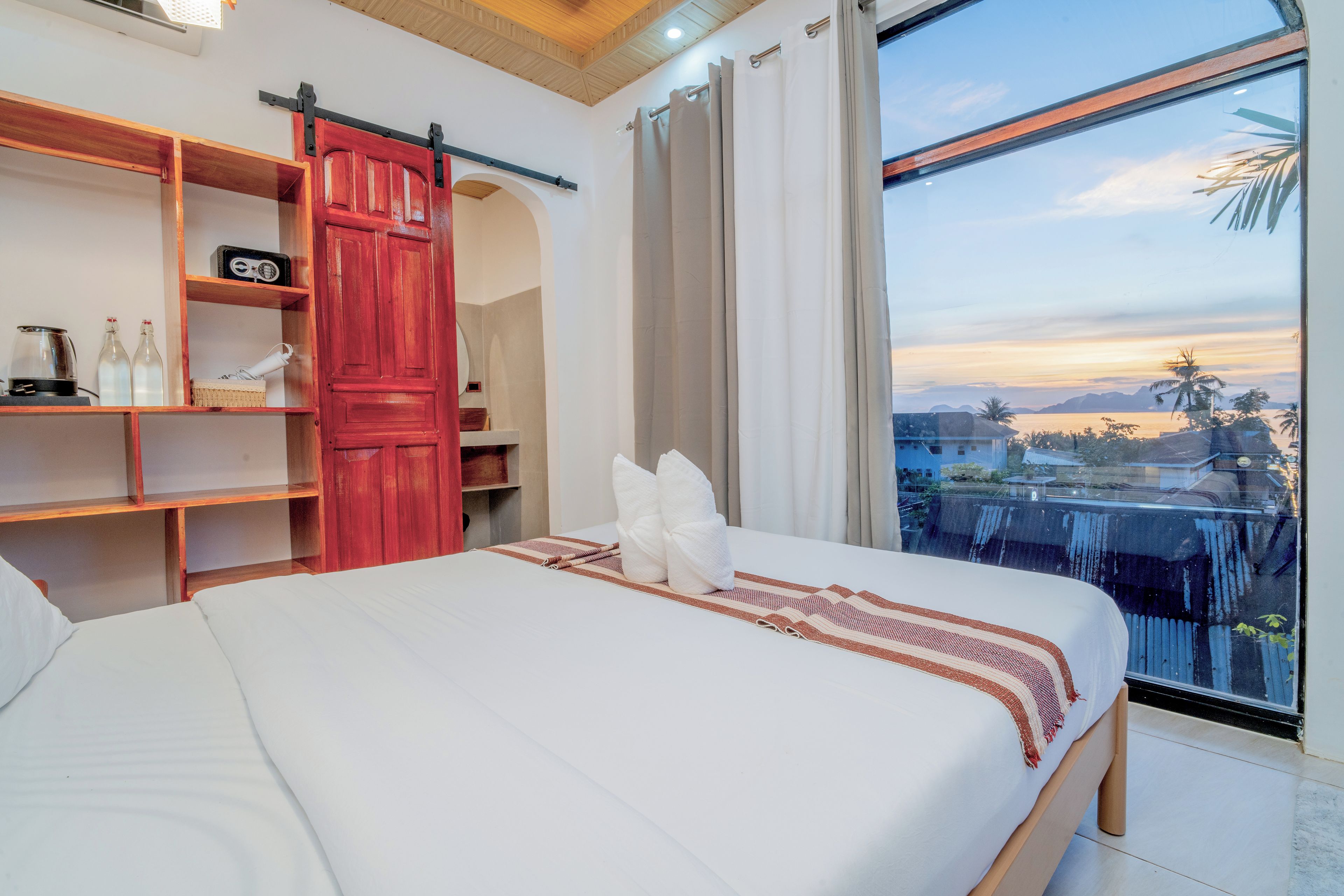 Ett hotellrum med en säng, en hylla i trä med flaskor, en röd dörr och ett stort fönster med utsikt över palmer och berg.